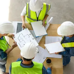 Zespół w kaskach i kamizelkach omawia plany domu. Trwa prowadzenie budowy.