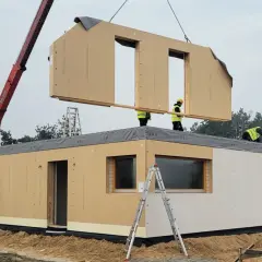 Budowa domu z gotowych elementów. Dźwig podnosi moduł dachu, pracownicy na konstrukcji montują kolejny element.