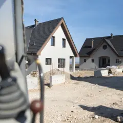 Budowa dwóch domów gospodarczych w słoneczny dzień. Widać fragment maszyny budowlanej na pierwszym planie.