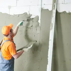 Murarz w pomarańczowej czapce i ogrodniczkach nakłada tynk na ścianę.