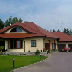 Elegancki dom w stylu włoskim z czerwoną dachówką, otoczony zielenią. Na podjeździe stoi czerwony samochód.