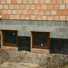 Widok na fragment ściany budynku z cegły klinkierowej, szarego betonu i izolacji przeciwwilgociowej. Dwa małe okna z brązowymi ramami. Widoczny poziom posadowienia budynku.