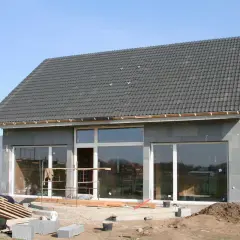 Dom w stanie surowym zamkniętym, z dachem i oknami. Widać materiały budowlane i pracującego człowieka.