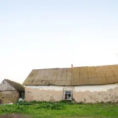 Dwa stare budynki gospodarcze na zgłoszenie, jeden z kamienia i drewna, drugi z tynkiem odpadającym od ścian, stoją na tle jasnego nieba.