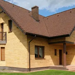 Jasny, ceglany dom z brązowym dachem i kominem. Widoczny jest cokół z ciemniejszej cegły, a także balkon i ganek.