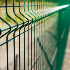Zielone ogrodzenie z siatki bez podmurówki, z charakterystycznymi przetłoczeniami, biegnie wzdłuż piaszczystej ścieżki.