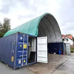 Tymczasowa hala z kontenerów morskich, z zielonym, łukowatym dachem, chroni sprzęt przed deszczem.