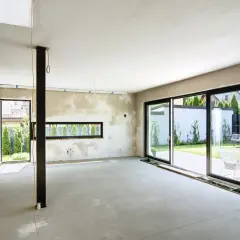 Wnętrze w trakcie budowy, ściany pokryte tynkami gipsowo wapienne, betonowa podłoga, duże okna z widokiem na ogród.