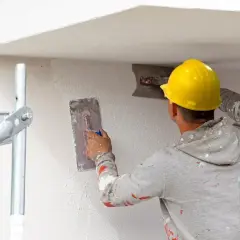Budowlaniec w żółtym kasku tynkuje ścianę za pomocą kielni.