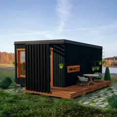 Nowoczesne domy z kontenerów, projekty i ceny. Czarny, minimalistyczny moduł mieszkalny z drewnianym tarasem i meblami ogrodowymi, otoczony zielenią i jeziorem.