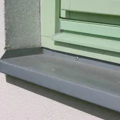 Szary parapet zewnętrzny zamontowany przy oknie z zielonymi okiennicami. Idealny moment, kiedy montować parapety zewnętrzne, to po ociepleniu i otynkowaniu elewacji.