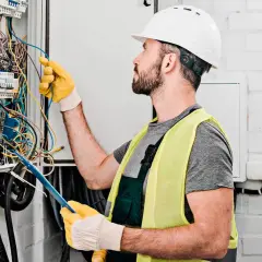 Elektryk w kasku i kamizelce odblaskowej pracuje przy skrzynce z przewodami. Dokładna wycena koszt instalacji elektrycznej wymaga precyzji.