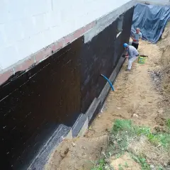 Dwóch mężczyzn wykonuje hydroizolację fundamentów czarną masą KMB.