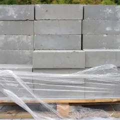 Stos szarych bloczków betonowych na palecie, owiniętych folią. Poznaj bloczek betonowy wymiary dla swojej budowy.