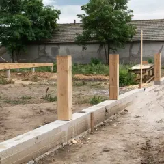 Solidny fundament pod ogrodzenie z betonowym murem i drewnianymi słupkami, w tle budowa domu.