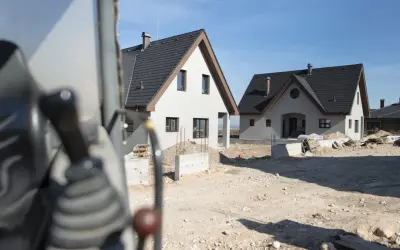 Budowa dwóch domów gospodarczych w słoneczny dzień. Widać fragment maszyny budowlanej na pierwszym planie.