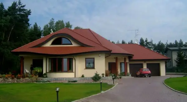 Elegancki dom w stylu włoskim z czerwoną dachówką, otoczony zielenią. Na podjeździe stoi czerwony samochód.