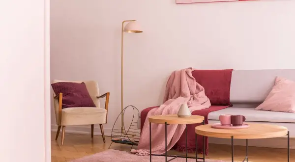 Przytulny salon z fotelem w kolorze ecru i amarantową poduszką. Na kanapie narzuta w tym samym amarantowym kolorze.