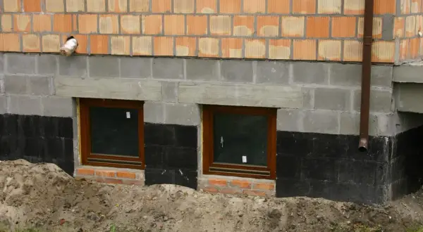 Widok na fragment ściany budynku z cegły klinkierowej, szarego betonu i izolacji przeciwwilgociowej. Dwa małe okna z brązowymi ramami. Widoczny poziom posadowienia budynku.