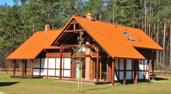 Urokliwy dom szachulcowy z pomarańczowym dachem, otoczony zielonym trawnikiem i lasem sosnowym.