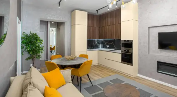Jasny salon z sofą i żółtymi poduszkami, okrągły stół z krzesłami, kuchnia w kolorze wanilia i drewna, telewizor.