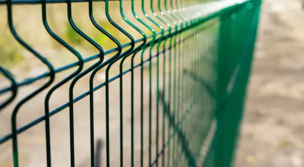 Zielone ogrodzenie z siatki bez podmurówki, z charakterystycznymi przetłoczeniami, biegnie wzdłuż piaszczystej ścieżki.