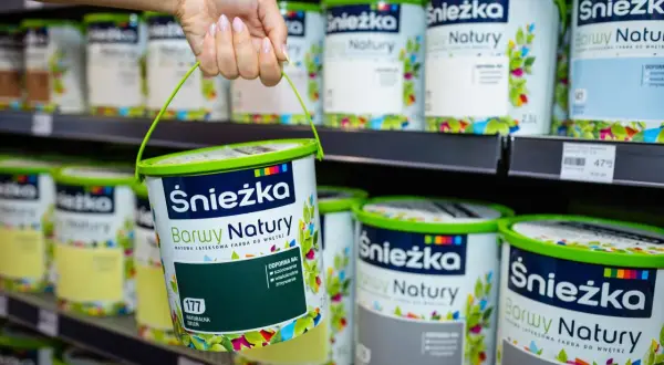 Ręka trzyma wiaderko z zieloną farbą Śnieżka Barwy Natury. Na półce widać wiele innych puszek tych farb.