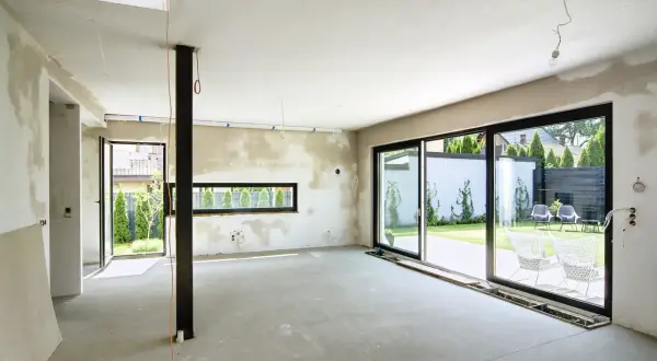 Wnętrze w trakcie budowy, ściany pokryte tynkami gipsowo wapienne, betonowa podłoga, duże okna z widokiem na ogród.