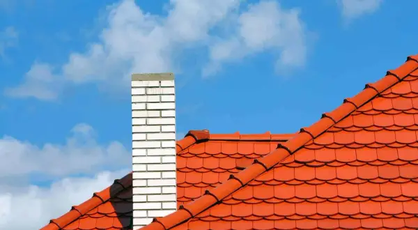 Pomarańczowy dach wielospadowy z ceglanym kominem na tle błękitnego nieba z chmurami.