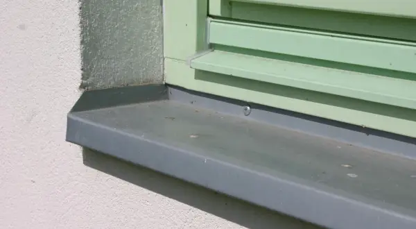 Szary parapet zewnętrzny zamontowany przy oknie z zielonymi okiennicami. Idealny moment, kiedy montować parapety zewnętrzne, to po ociepleniu i otynkowaniu elewacji.