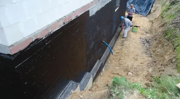 Dwóch mężczyzn wykonuje hydroizolację fundamentów czarną masą KMB.