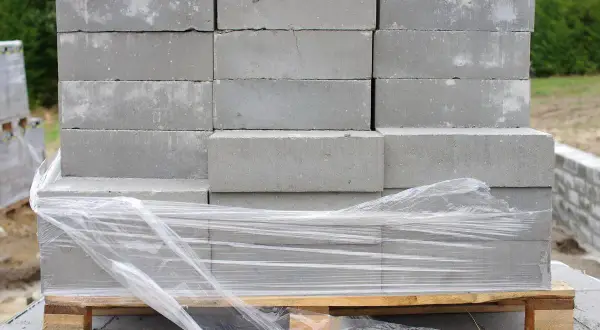 Stos szarych bloczków betonowych na palecie, owiniętych folią. Poznaj bloczek betonowy wymiary dla swojej budowy.