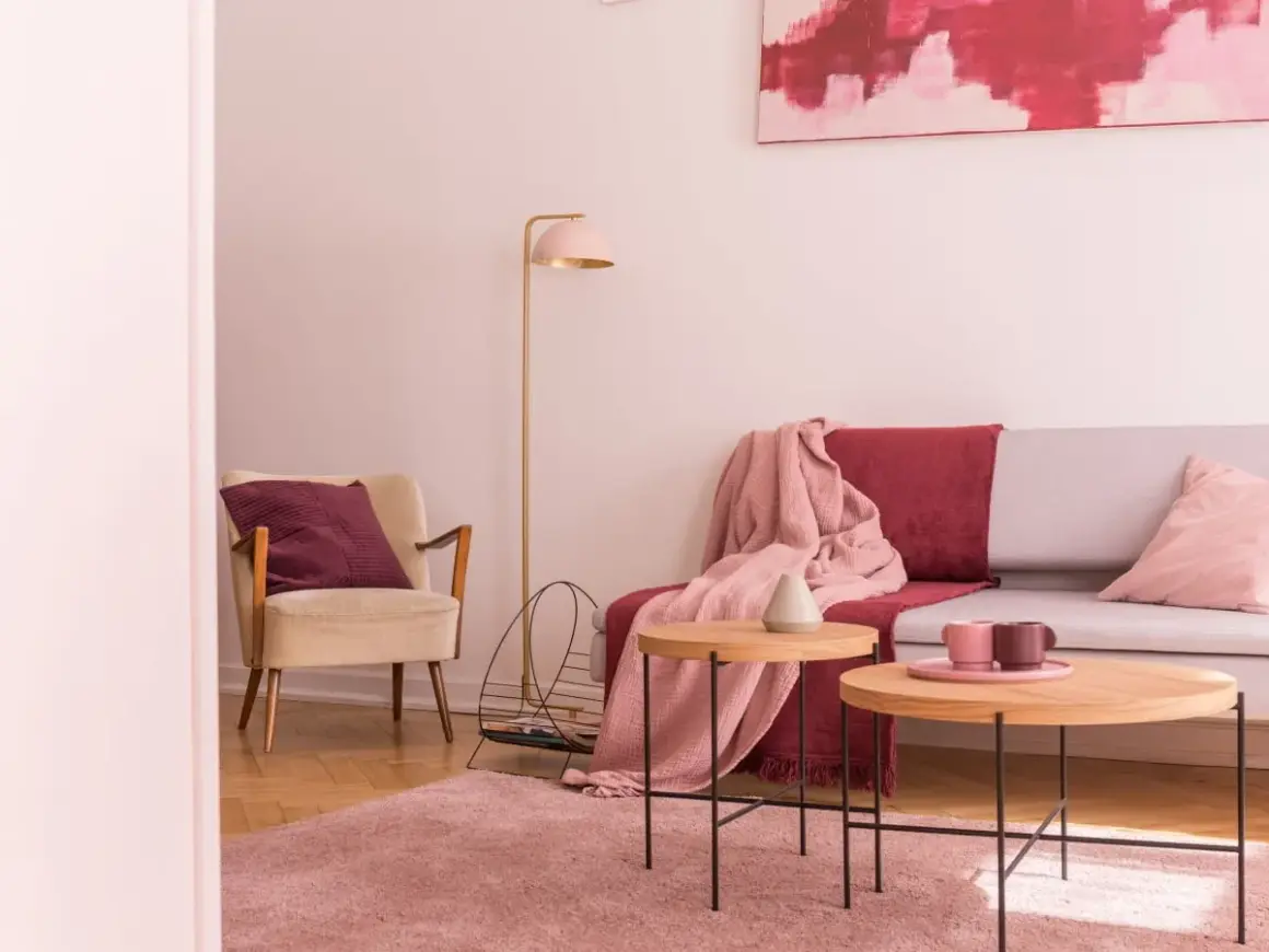 Przytulny salon z fotelem w kolorze ecru i amarantową poduszką. Na kanapie narzuta w tym samym amarantowym kolorze.