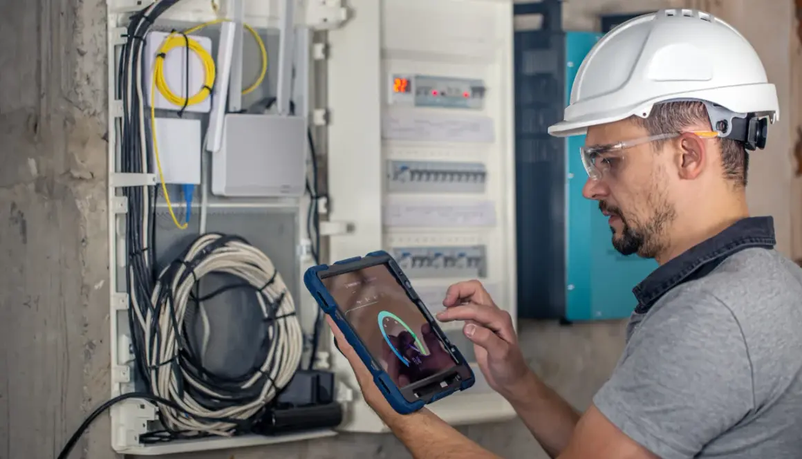 Technik w kasku i okularach ochronnych wykonuje przegląd instalacji elektrycznej, korzystając z tabletu z aplikacją diagnostyczną.