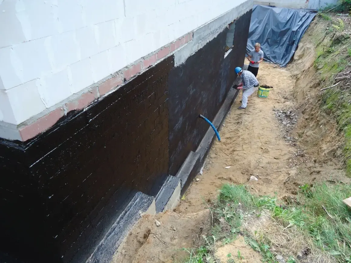 Dwóch mężczyzn wykonuje hydroizolację fundamentów czarną masą KMB.