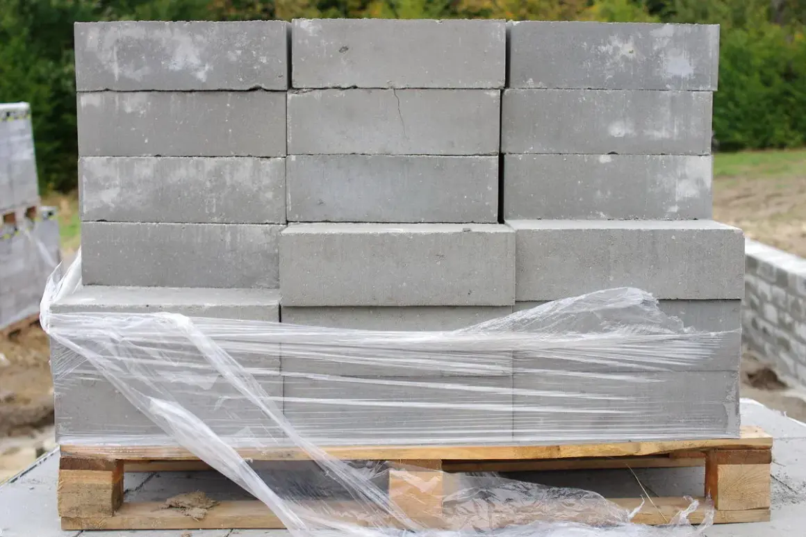 Stos szarych bloczków betonowych na palecie, owiniętych folią. Poznaj bloczek betonowy wymiary dla swojej budowy.