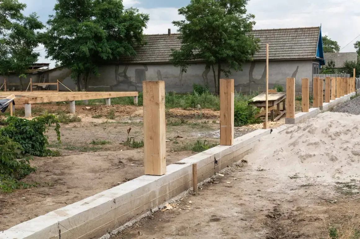 Solidny fundament pod ogrodzenie z betonowym murem i drewnianymi słupkami, w tle budowa domu.