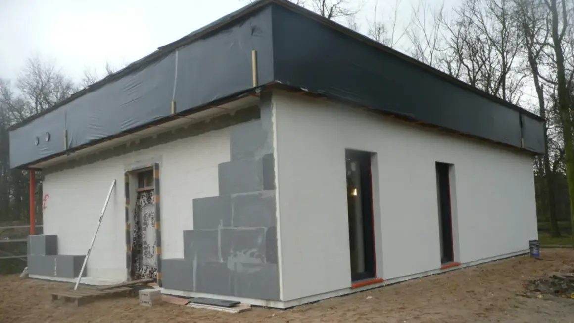 Budowa domu ze styropianu w toku. Ściany z bloczków, częściowo otynkowane, dach przykryty czarną folią.
