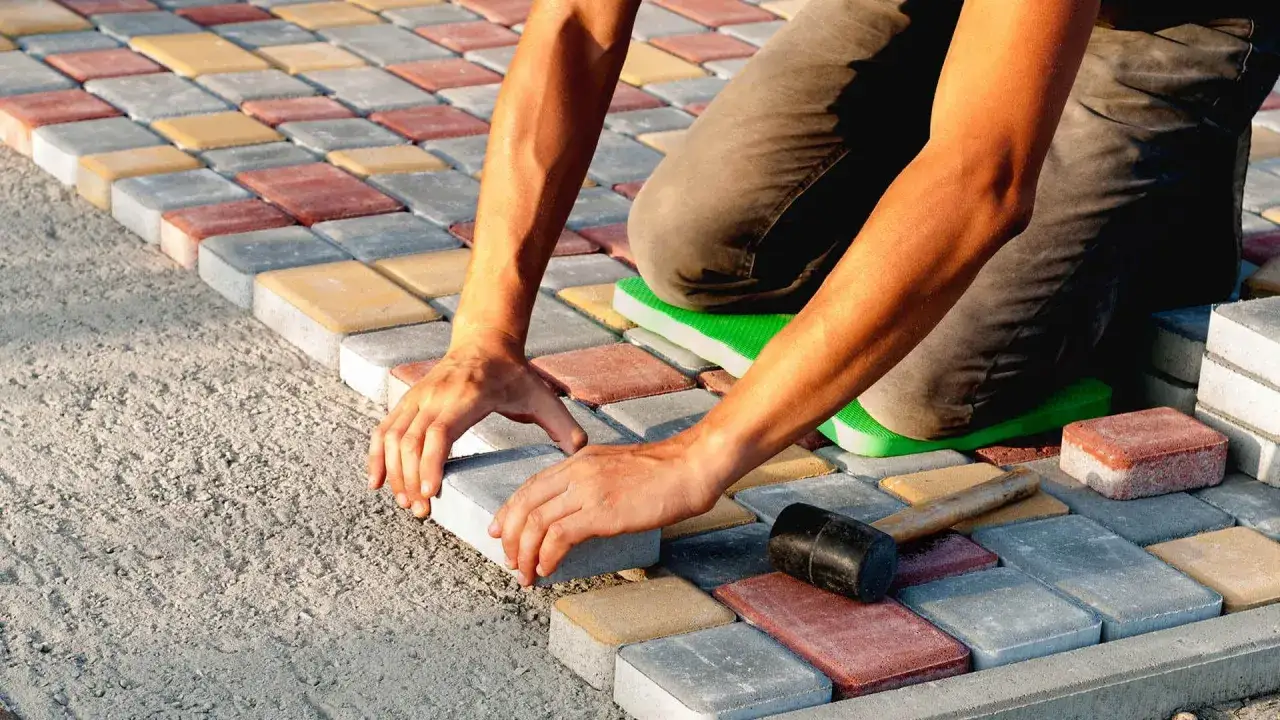 Układanie kolorowej kostki brukowej na podsypce. Widać ręce pracownika i młotek. Potrzebna wiedza, ile suchego betonu pod kostkę.