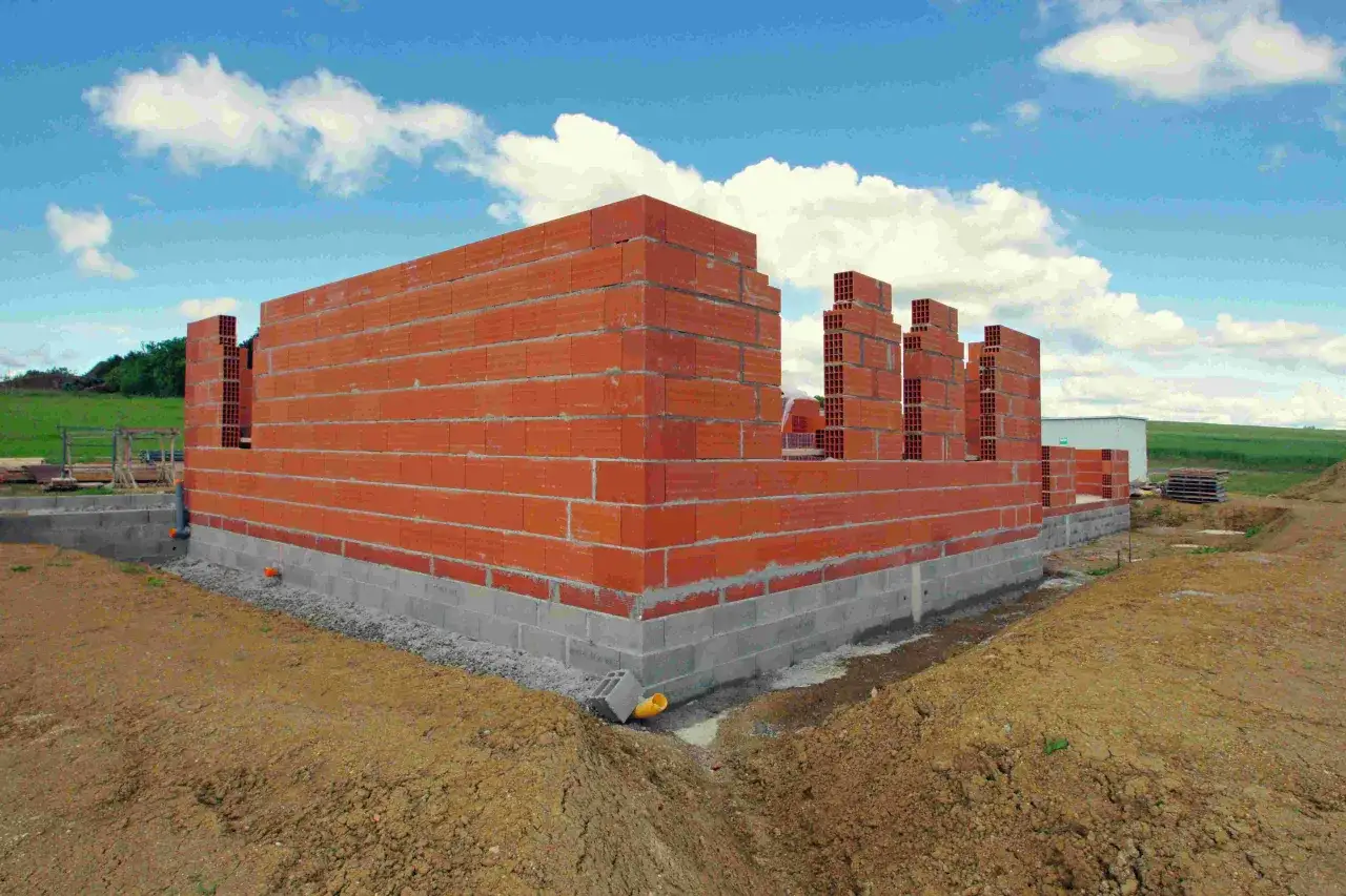 Budowa domu w toku: czerwone ściany z pustaków piętrzą się na betonowym fundamencie. Widać zarys pomieszczeń, a niebo błękitne z chmurami.