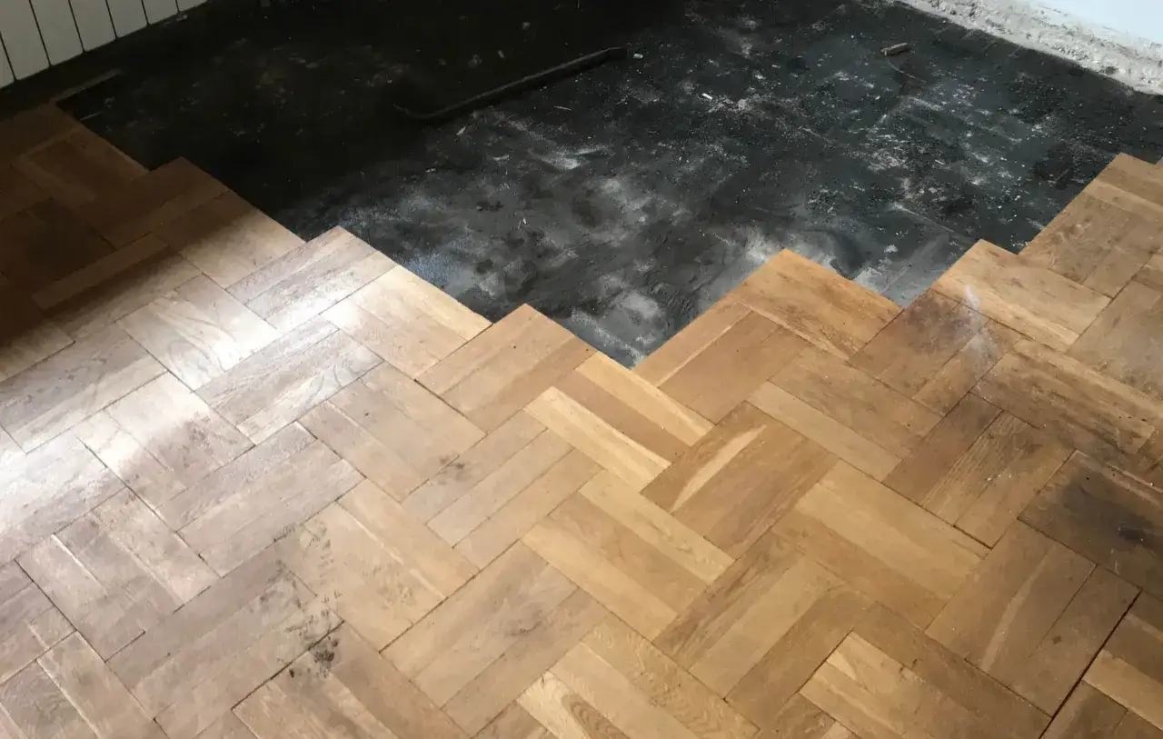 Układanie parkietu w jodełkę. Część podłogi jest już gotowa, a obok widać przygotowany podkład. subit co to? To klej do parkietu.