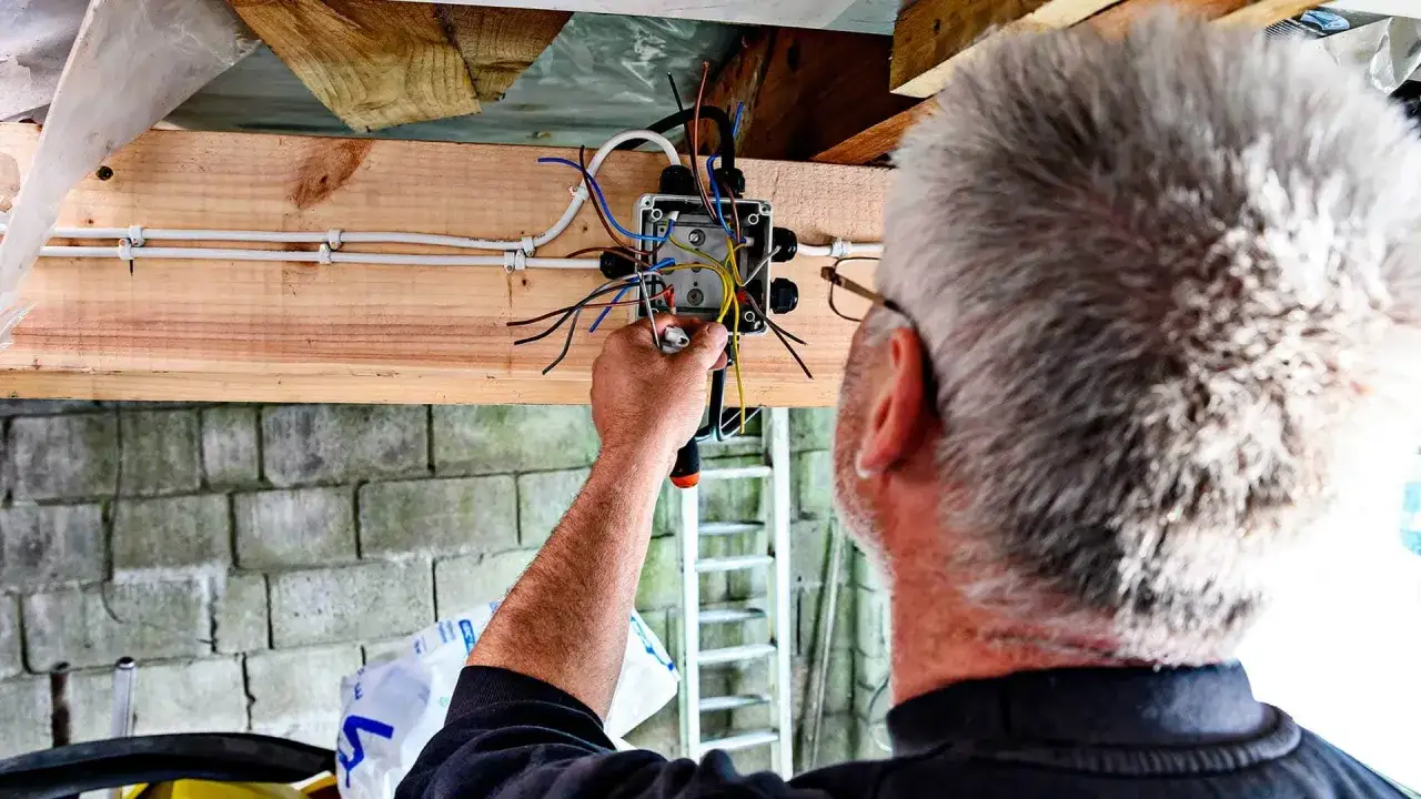 Mężczyzna z siwymi włosami pracuje nad instalacją elektryczną w garażu, łącząc przewody w puszce.