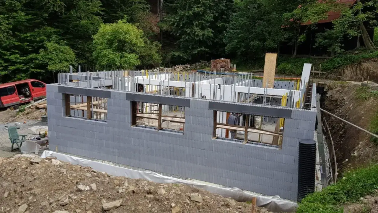 Budowa domu z piwnicą w lesie. Widać szary betonowy fundament, okna i zbrojenie. W tle czerwony van i drzewa.