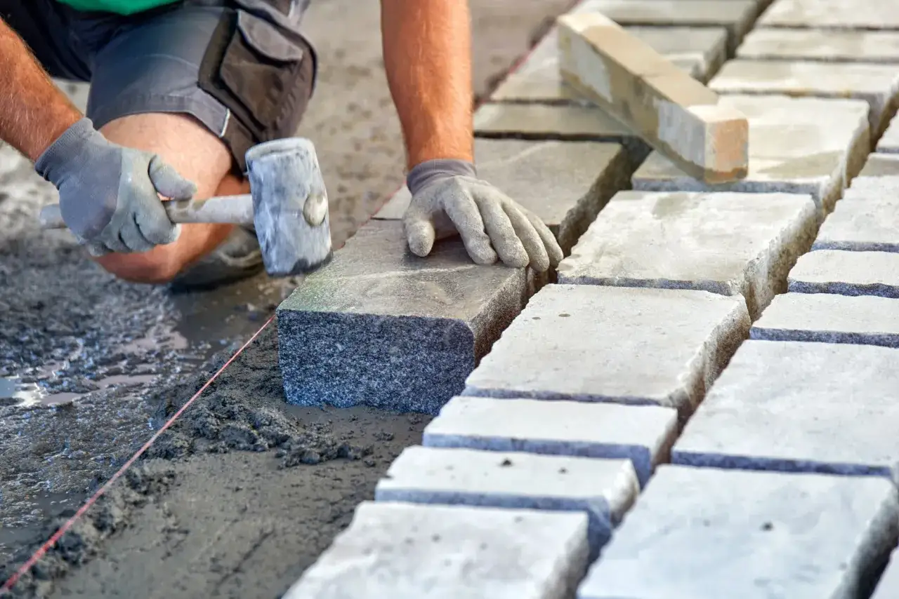 Układanie kostki brukowej na podsypce. Ręce w rękawiczkach kładą kamień, używając gumowego młotka. Widać suchy beton proporcje.