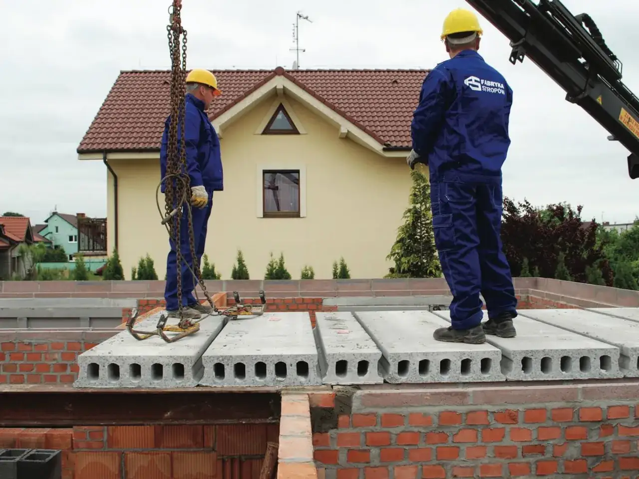 Dwaj pracownicy w żółtych kaskach i niebieskich kombinezonach montują płyty stropowe. Ich praca wymaga precyzji, niczym przy tworzeniu filigranowych ozdób.