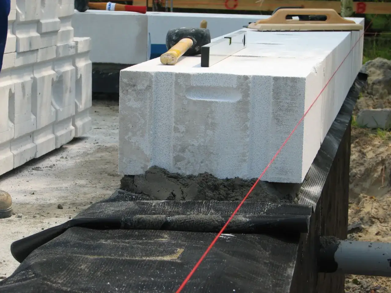Budowa ścian fundamentowych z bloczków betonowych. Na budowie widać młotek, poziomicę i sznur murarski.
