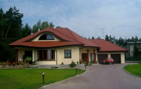 Elegancki dom w stylu włoskim z czerwoną dachówką, otoczony zielenią. Na podjeździe stoi czerwony samochód.