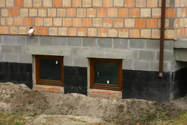 Widok na fragment ściany budynku z cegły klinkierowej, szarego betonu i izolacji przeciwwilgociowej. Dwa małe okna z brązowymi ramami. Widoczny poziom posadowienia budynku.