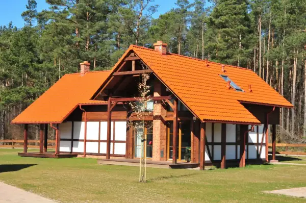 Urokliwy dom szachulcowy z pomarańczowym dachem, otoczony zielonym trawnikiem i lasem sosnowym.
