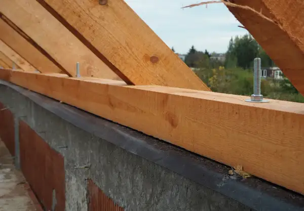 Drewniana murłata z widocznymi wymiarami, przykręcona do betonowego wieńca. W tle drzewa i domy.
